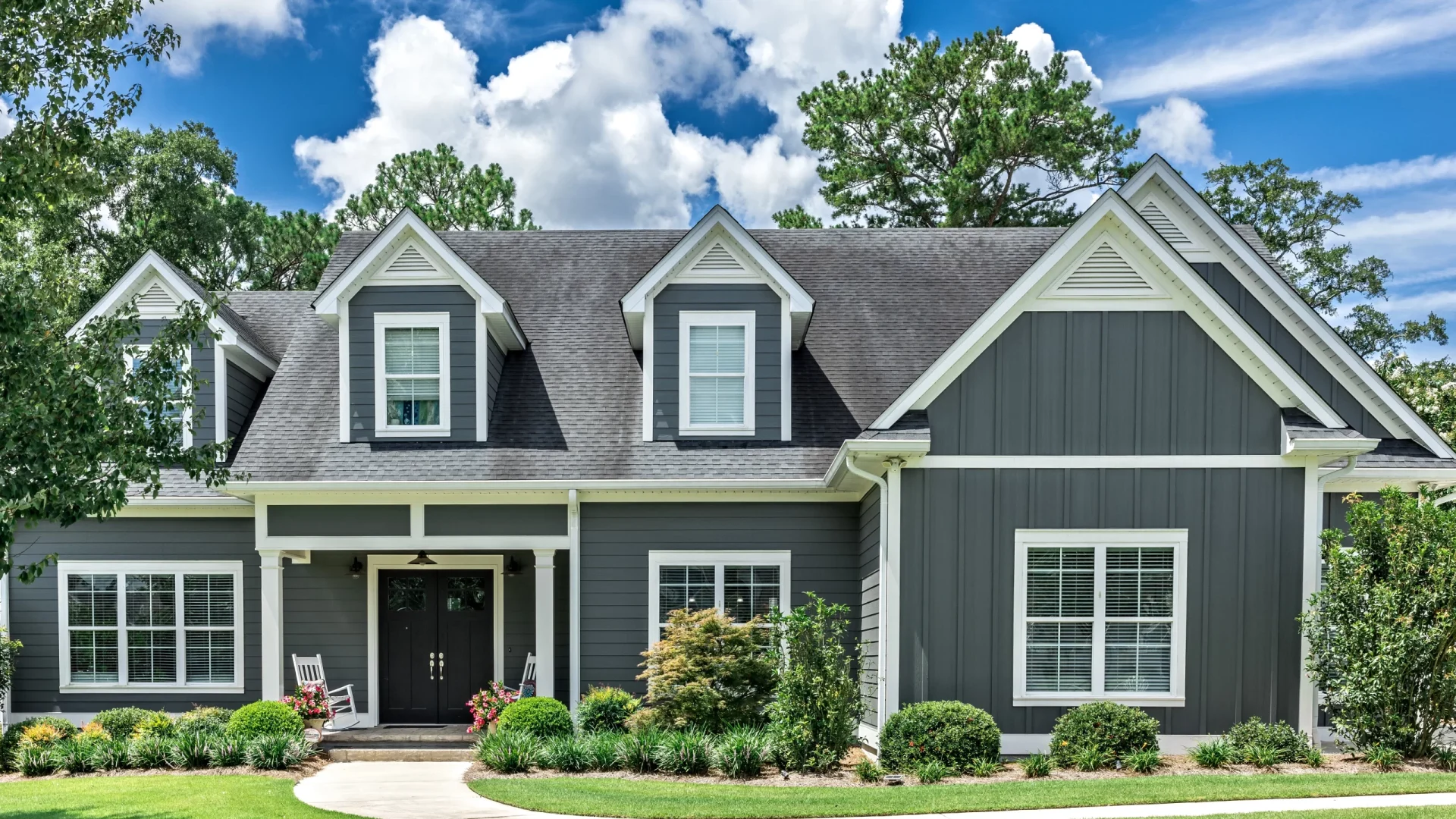Front of a gray-sided house with white trim, black double front entry doors, gray asphalt shingle roofing, and three dormers